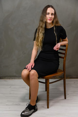 Vertical photo of a young pretty woman in a black dress on a gray background in the studio. A model with long hair is sitting on a chair.