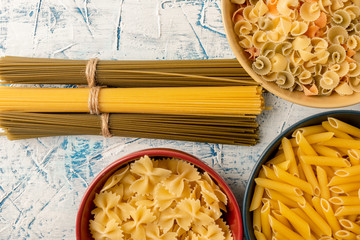 Spaghetti in bunches and various pastas in multicolored ceramic bowls on a blue and white textured background. Long-term storage products