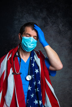 Distraught male doctor with hand on head, wearing blue hospital scrubs with face mask & stethoscope, holding the American flag close to his chest, against a dark studio background.