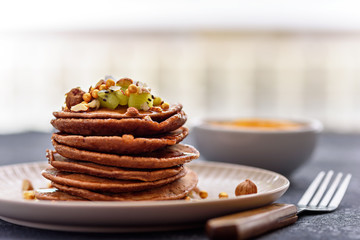 stack chocolate pancakes with kiwi, hazelnut, maple syrup on plate with fork on kitchen table