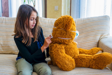 Beautiful little girl in a black jacket playing with her brown teddy bear who has put a mask to protect him from the coronavirus on the white sofa at home