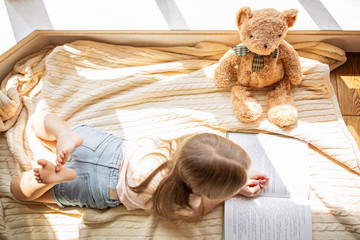 Cute little caucasian girl in casual clothes reading a book with stuffed teddy bear toy and smiling while lying on a floor near window in the room. Stay at home during coronavirus covid-19 pandemic