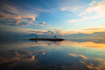 Sunrise seascape. Mountains and Agung volcano. Traditional gazebos on an artificial island in the ocean. Water reflection. Sanur beach, Bali, Indonesia.