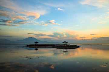 Fototapeta na wymiar Sunrise seascape. Mountains and Agung volcano. Traditional gazebos on an artificial island in the ocean. Water reflection. Sanur beach, Bali, Indonesia.