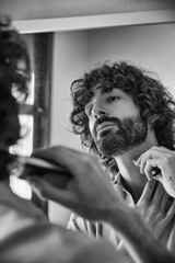 Handsome young man looking in the mirror while fixing his beard. Lifestyle