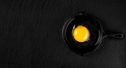 Fresh raw chicken egg broken in a cast iron or steel pan, on a dark stone background. Top view of a bright yolk, copy space for text or recipe. Breakfast or lunch concept