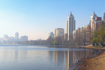 Obraz premium Scenic spring landscape view of Dnieper River and empty beach in modern Obolon district. Embankment with skyscrapers in morning smog in the background. Buildings reflected in the water. Kyiv, Ukraine