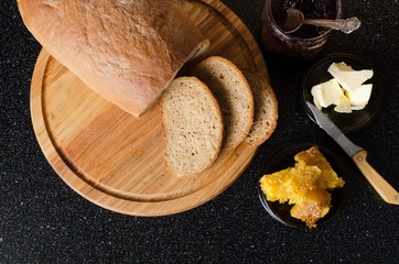 Top view of wooden cutting board and tasty rye bread on it, honey, butter, jam on the black kithcen table.Concept of delicious and sweet breakfast in the morning