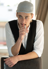 A young man in a cap and vest smokes by the window. Israel