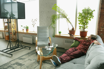 Watching TV lying on sofa. Caucasian man staying at home during quarantine because of coronavirus, COVID-19 spreading. Trying to spent time fun. Concept of healthcare and medicine, isolation.