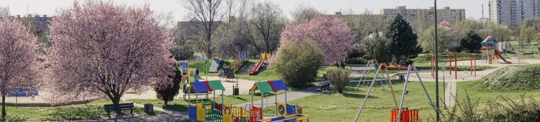 wiosenna panorama z placem zabawa dla dzieci, kwitnące drzewa i krzewy, przyrządy do zabaw i ćwiczeń, pusty plac w okresie pandemii - obrazy, fototapety, plakaty