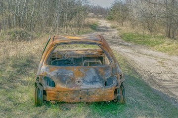 spalony porzucony samochód na leśnej drodze, wiosna odsłania zimowe śmieci, wiosenny las bez liści, recykling samochodów