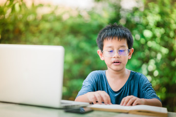 The asian boy wearing the glasses reading book in the garden,.The boy concentrate for read the book by the laptop beside him, The boy learning from home by laptop