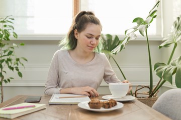Obraz na płótnie Canvas Teen girl eating cupcakes, drinking tea, sitting at table at home