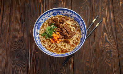 Chinese beef noodle soup in traditional blue bowl, fresh herbs and sliced carrot, pair of chopsticks on dark wooden background and copy space. Asian food concept top view