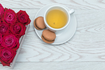 Obraz na płótnie Canvas Red roses in gift box on white wooden table. Cup of tea and chocolate macaroons on a plate. Top view. Sweet dessert as a present on a table, minimal style.