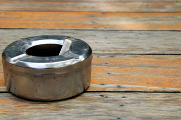 Round shape stainless ashtray is on light brown wooden floor and a cigarette in it, Thailand. 