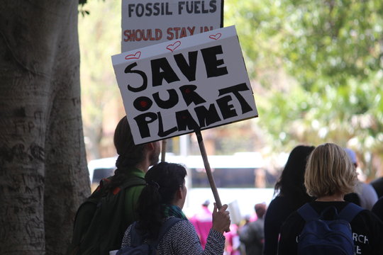 People At A Climate Change Protest. They Are Holding Up Signs. One Of Them Reads 