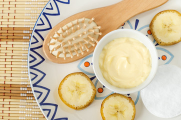 Diy banana and yogurt mask (face cream) in the small white bowl, wooden hair brush, cotton pads and banana slices. Natural homemade hair treatment and zero waste concept. Top view, copy space