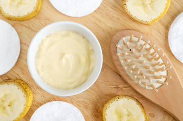 Diy banana and yogurt mask (face cream) in the small white bowl, wooden hair brush, cotton pads and banana slices. Natural homemade hair treatment and zero waste concept. Top view, copy space