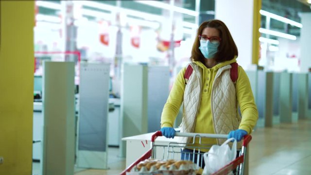 Woman in mask walking along cash registers with shopping cart full of food