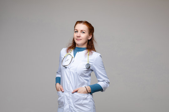 Stock photo portrait of a red-haired professional doctor in white coat with stethoscope around her neck posing with her hands in pockets. Isolate on grey background. Studio shot.