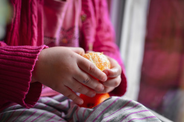 The baby is sitting on the window during quarantine. The child is sitting at home. Girl holding tangerines in her hands.