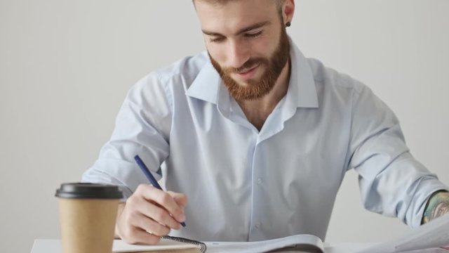 An attractive young bearded man wearing a shirt is writing something sitting at the table over a white wall