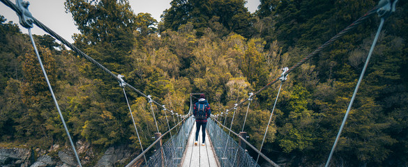 Hokitika Gorge, New Zealand, October 7, 2019: Beautiful panorama of a European man with a photographic backpack crossing a suspension bridge surrounded by leafy trees