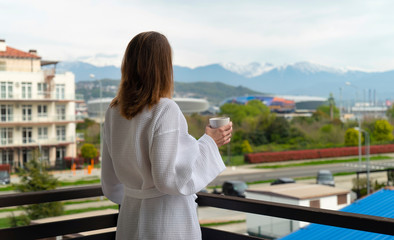 Fototapeta na wymiar Woman in a bathrobe on a balcony drinks coffee and looks at the street