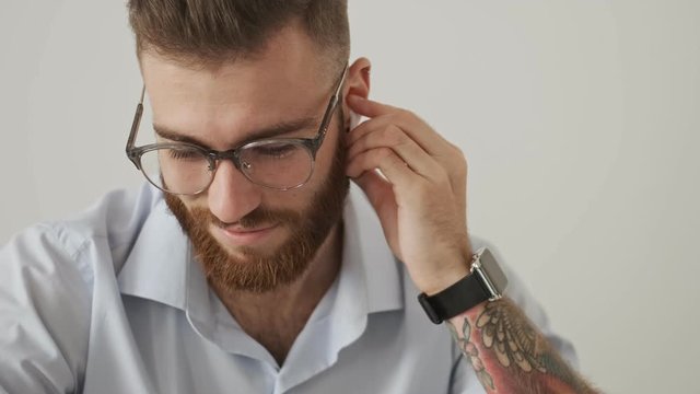 A positive bearded young businessman in eyeglasses is putting on his airpods over white wall