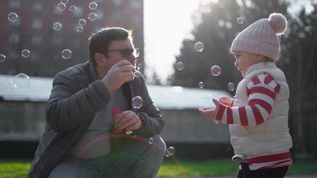 Caucasian man with his child blowing soap bubbles outside. Slow motion. Weekend activity with toddler, backyard