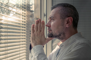 Man in isolation praying near a window blinds. Coronavirus outbreak. Man looks at a sunny street through the window blinds
