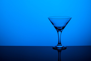 Glass goblet without wine on a thin leg stands on a mirror surface. Back light. Blue background. Copy space.