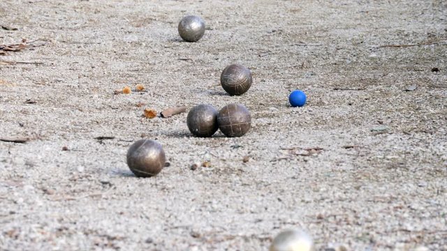 Game of petanque with a shining iron ball being thrown at a small blue plastic ball on a dirty ground full of pebbles and autumn leaves behind three iron balls sitting in the foreground