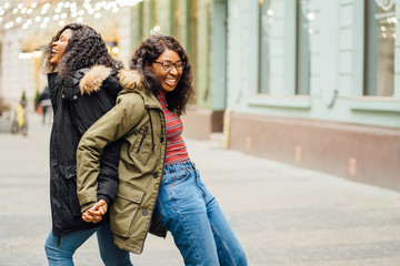 Portrait of two nigerian black women outdoor having fun hugging riding piggyback in cold weather. Two female student enjoying holiday in street. Happiness, fooling around, togetherness concept