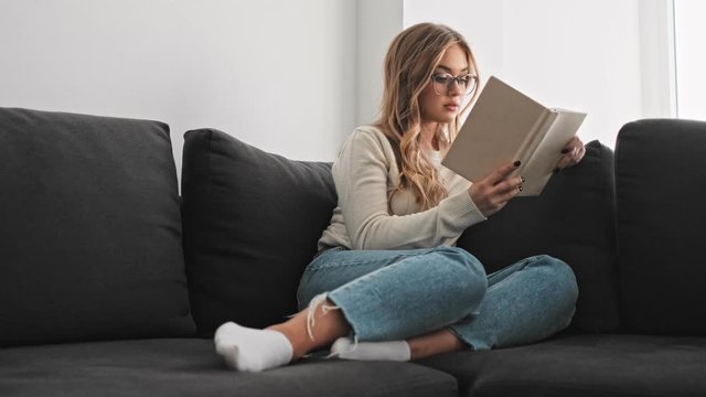 A smiling young woman is reading a book while sitting on the couch in the morning
