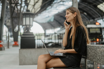 Girl is holding her smartphone making a call. Young blonde woman sitting on the stone bench and using her grey laptop for chatting with friends. She is waiting for the train at the railway station.