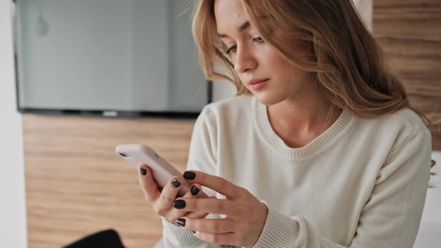 A pleased young blonde woman is using her phone in the kitchen in the morning