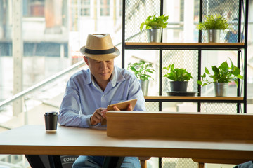 portrait middle aged Asian man in casual light blue shite and jeans pant and a cool hat sitting side coffee table bar using application on tablet with plant shelf with city urban building background.