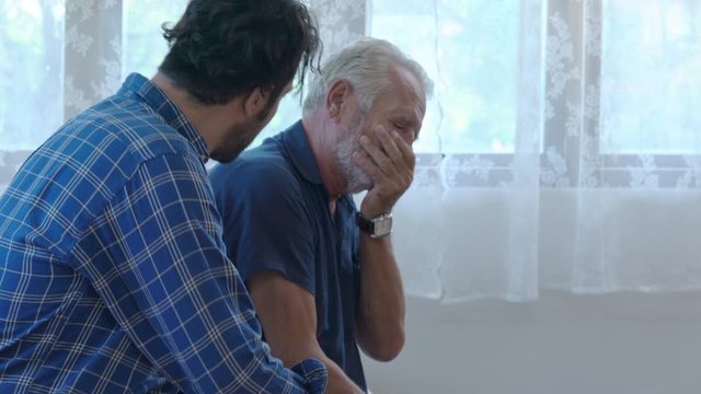 4K Handsome adult Caucasian man comforting his depressed senior father in the bedroom. Son hugging and consoling sadness old age dad with love. Mental health, healthcare and social problems concept.