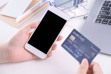 Woman holding smart mobile phone, concept of staying home online paying, electronic payment shopping on white table background with shop cart trolley, close up.