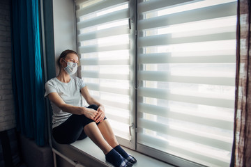 Side view young woman in wearing protective face mask sitting on windowsill at home, looking outside. Self-isolation, quarantine coronavirus pandemic prevention. Epidemic and prevention concept.