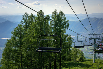 Ski lift in the mountains on sunny day against blue sky, white clouds, green hills and mountain lake. Mountain valley with cable car, view from top. Ski resort in summer.