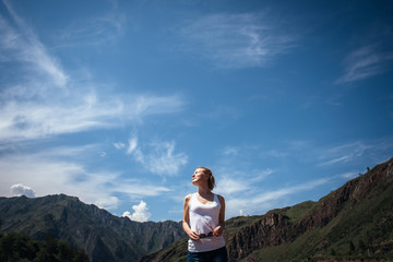 Naklejka na ściany i meble Happy female traveller in a white t-shirt against the beautiful mountains and blue sky on sunny day. Cheerful young girl enjoying a trip in the Altai mountains.