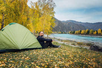 Young man sits on the grass near tent looking at the wildlife on autumn day in mountains. Hipster guy resting alone on bank of mountain river after trekking. Leisure tourism active lifestyle concept.