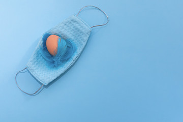 Easter quarantine flat lay of reusable face mask, feathers hand-painted egg on a blue background. Easter minimal creative greeting card. Copy space. Coronavirus.
