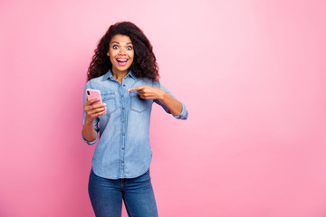 Look black firday. Addicted surprised afro american girl use smartphone point index finger impressed social network news wear denim jeans shirt isolated over pink color background