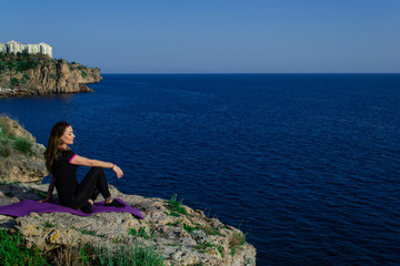 Naklejka na ściany i meble Beautiful young happy girl sitting on rock relaxing and looking to the blue sea
