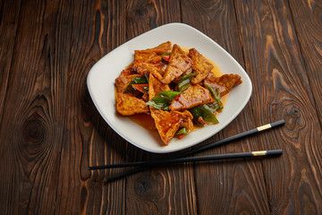 Homemade tofu stir fry with vegetables in hot sweet sauce and pair of chopsticks on dark wooden background and copy space. Asian food concept top view